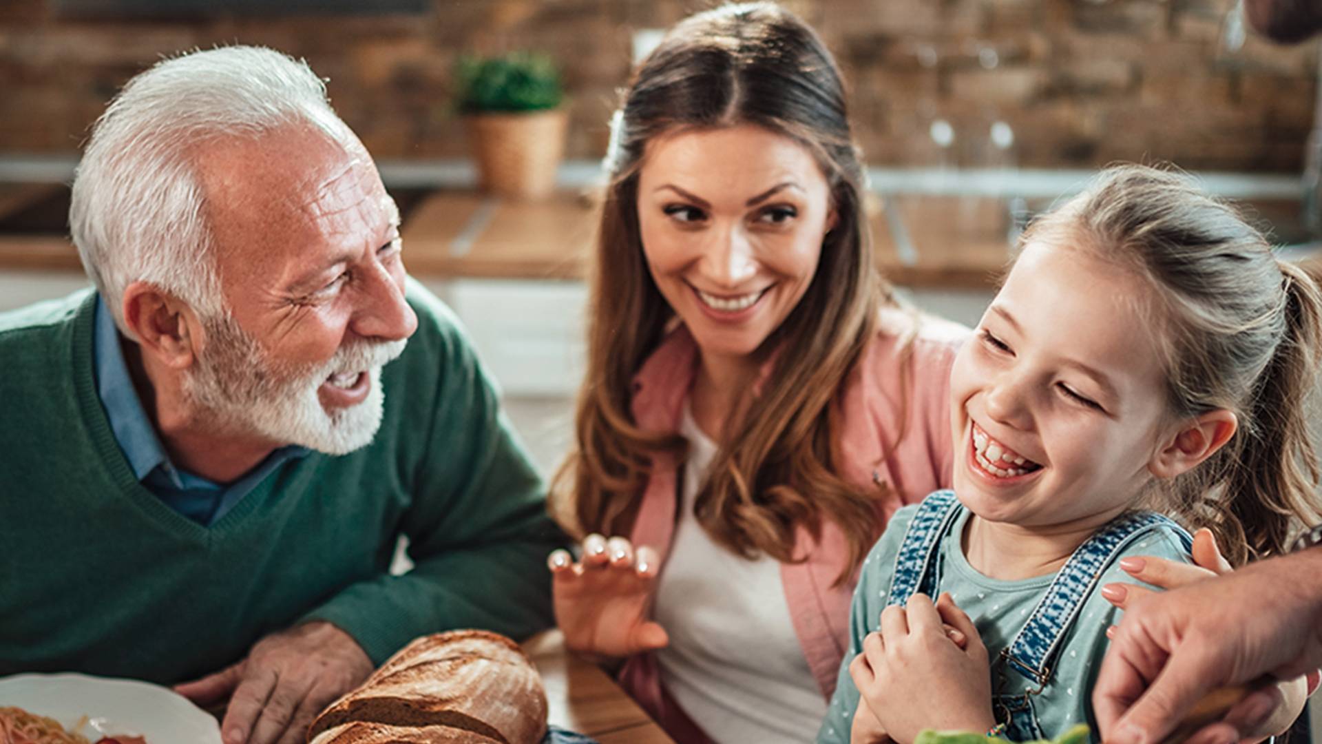 Un gruppo di persone sedute attorno a un tavolo da pranzo mentre condividono un pasto, con vari piatti tra cui pane, insalata, riso e un piatto di gamberi con verdure in una cucina accogliente.