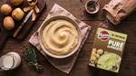 Ciotola di purè di patate accanto a una confezione di purè Pfanni e ingredienti freschi su un tavolo di legno.