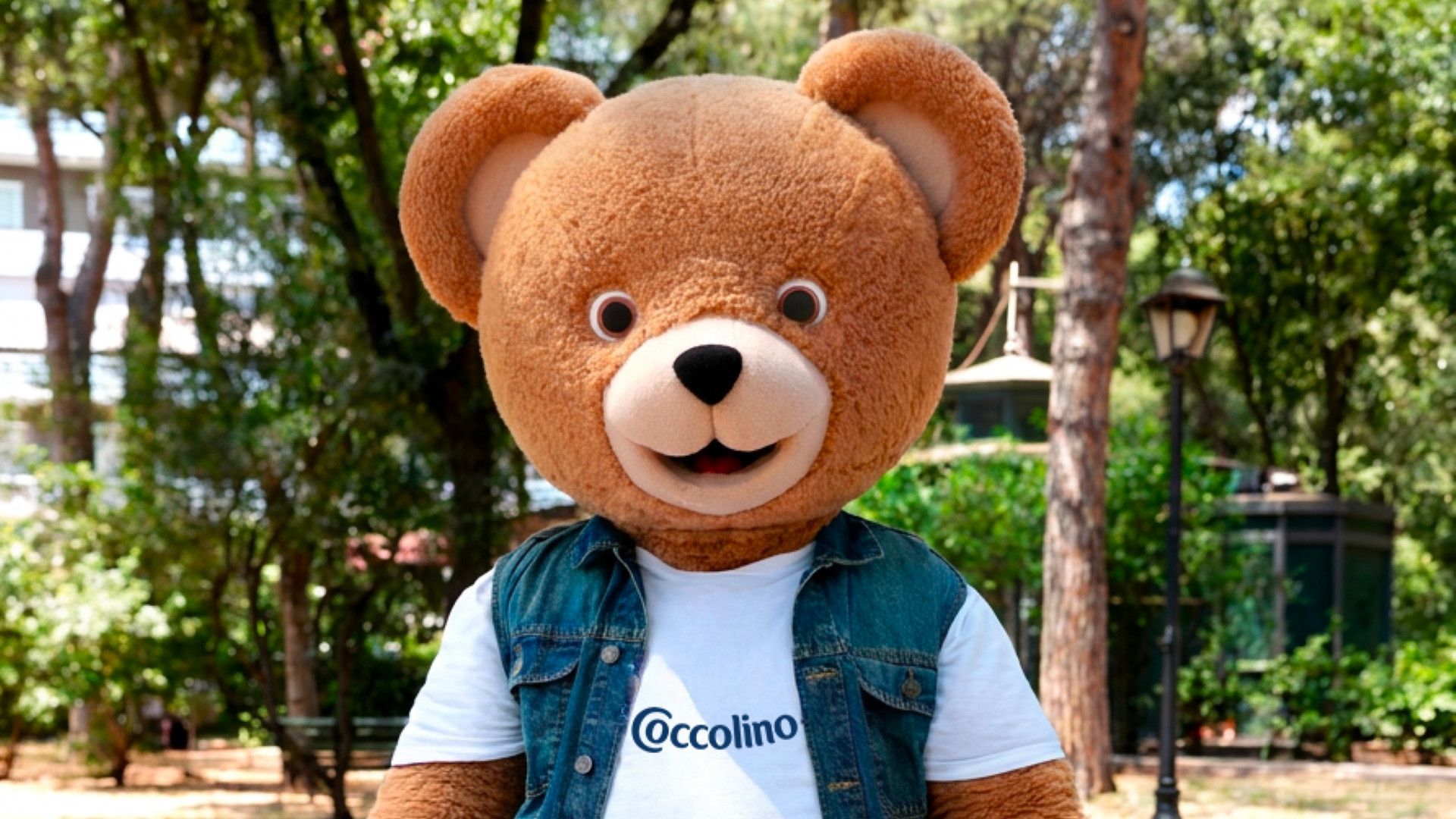 Mascotte orso Coccolino con T-shirt del marchio e gilet di jeans in un parco verdeggiante