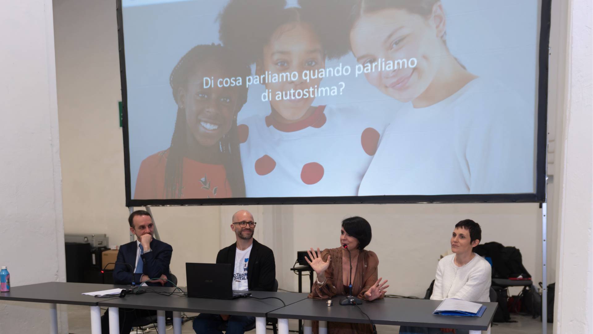 Seduti ad un tavolo ci sono degli speaker che parlano dei topic dell’evento con una presentazione alle spalle.